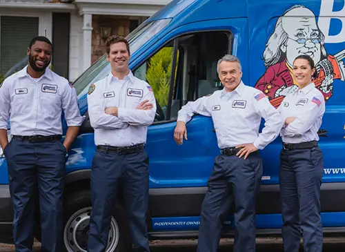 Four Benjamin Franklin Plumbing techs smiling at camera in front of a blue service vehicle